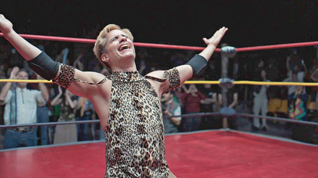 A wrestler with short hair stands in the center of a ring with his hands in the air. He's wearing a black and gold uniform.