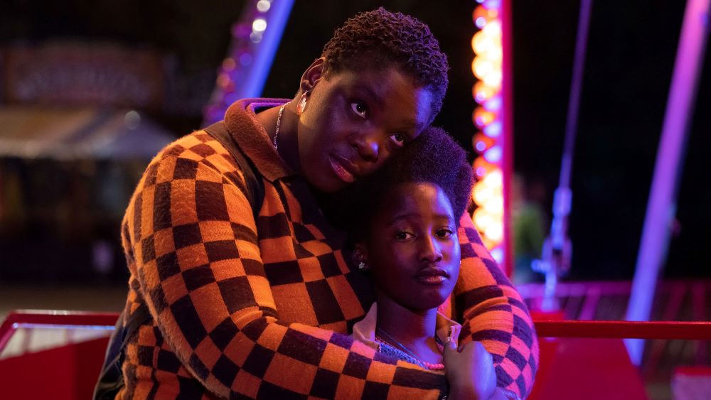 Black woman with closely cropped hair, in an orange and black checked shirt, stands, arms around, cheek touching the hair of a young Black Girl in a neat Afro, both with somber expressions