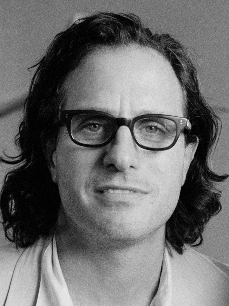 black and white photo of a white man with shoulder length hair looking at the camera with glasses
