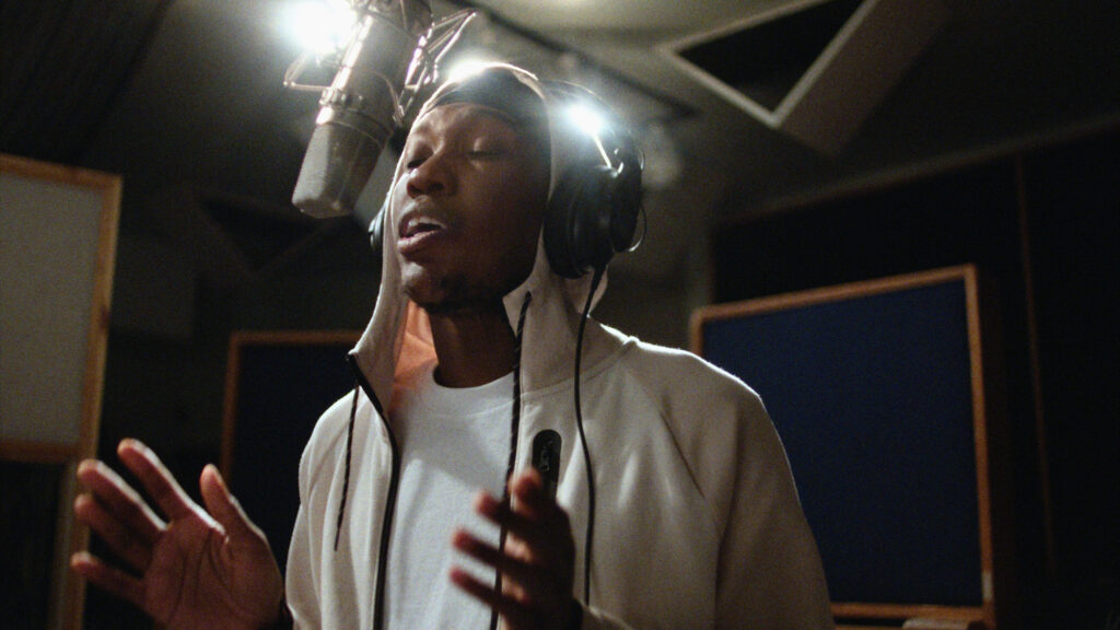 A film still from As We Speak that shows rap artist Kemba singing into a microphone in a recording studio.