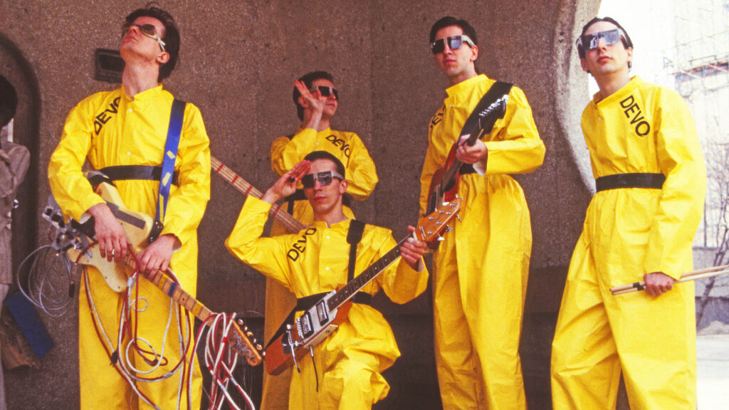 A film still from DEVO that shows the band Devo in matching yellow jumpsuits and sunglasses.