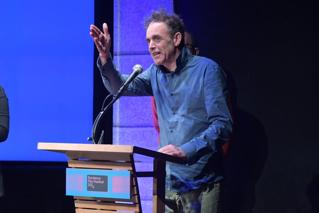 Man with rumpled hair gestures as he speaks into a microphone from a podium at the 2024 Sundance Film Festival