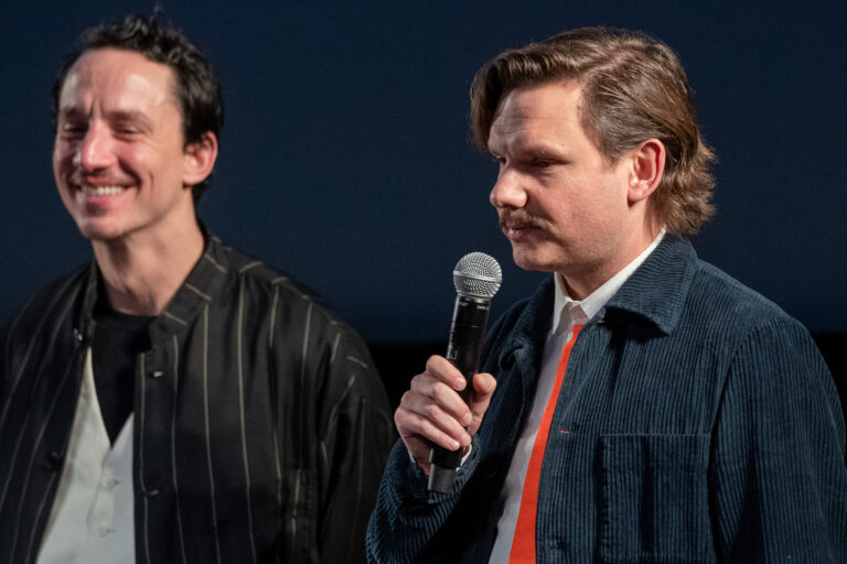 A laughing dark-haired man stands next to a man, with brown hair and mustache, holding a microphone.