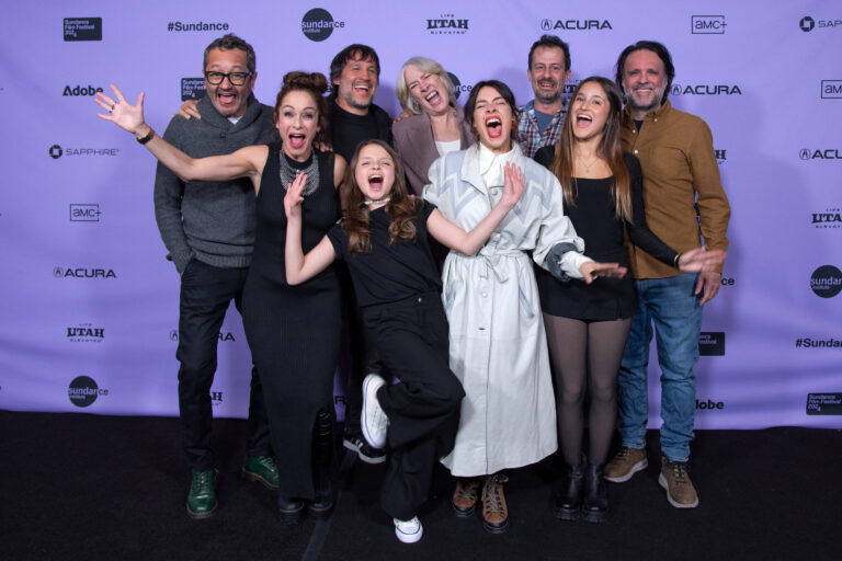 Group of men and women, of mixed ages, appear to scream and yell in jubilation before a 2024 Sundance Film Festival backdrop.