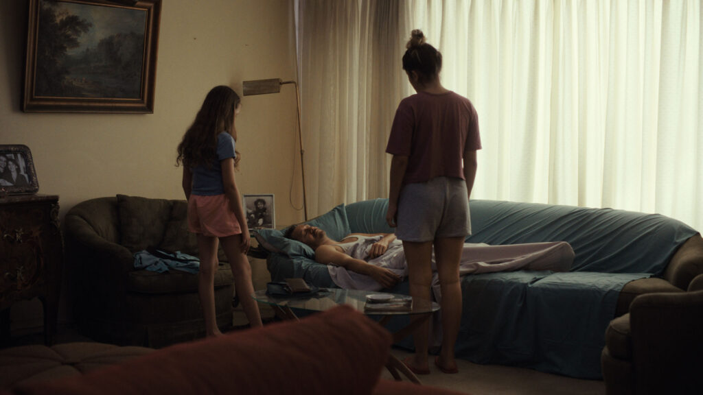 Reinas- Two girls stand over their sleeping father